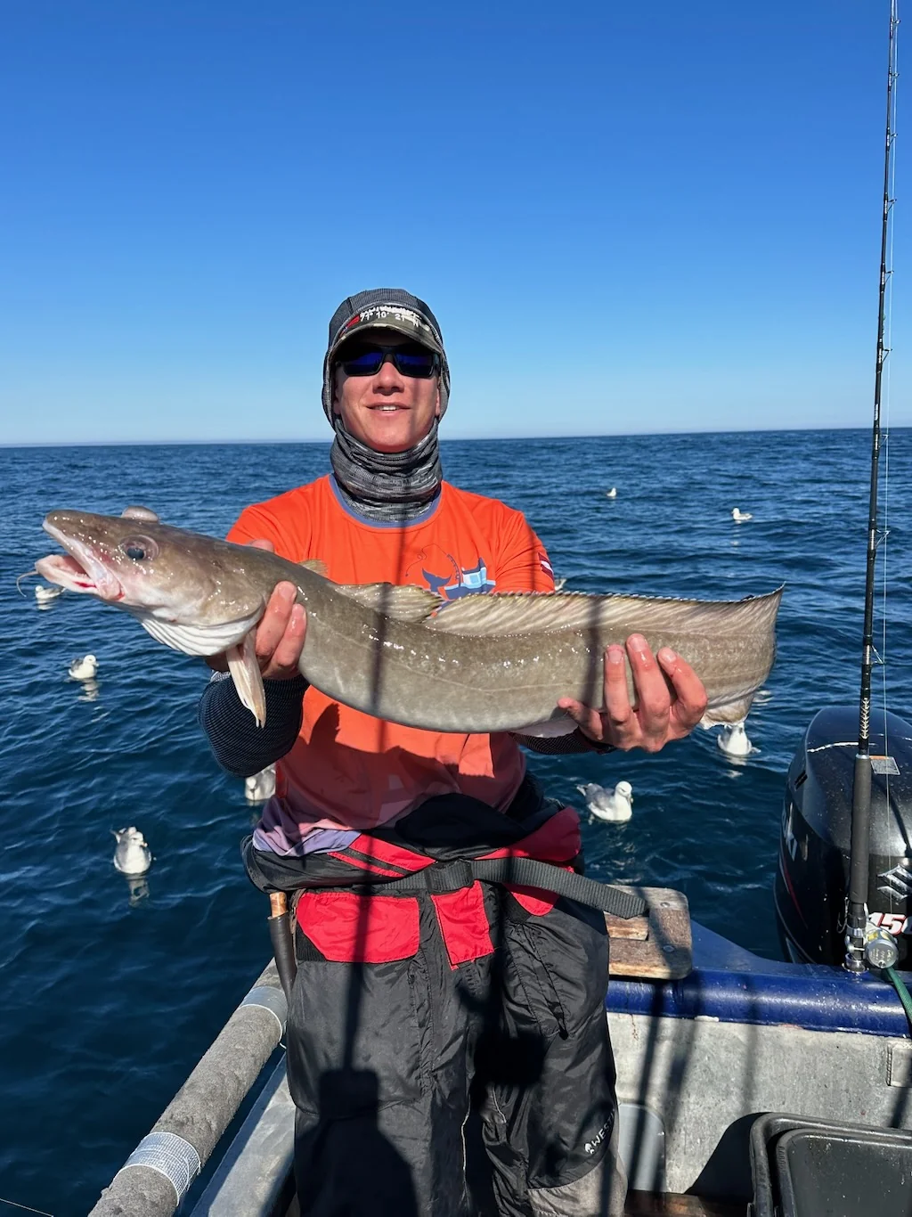 An angler in an orange shirt holding a fish on a boat, with blue sea behind.