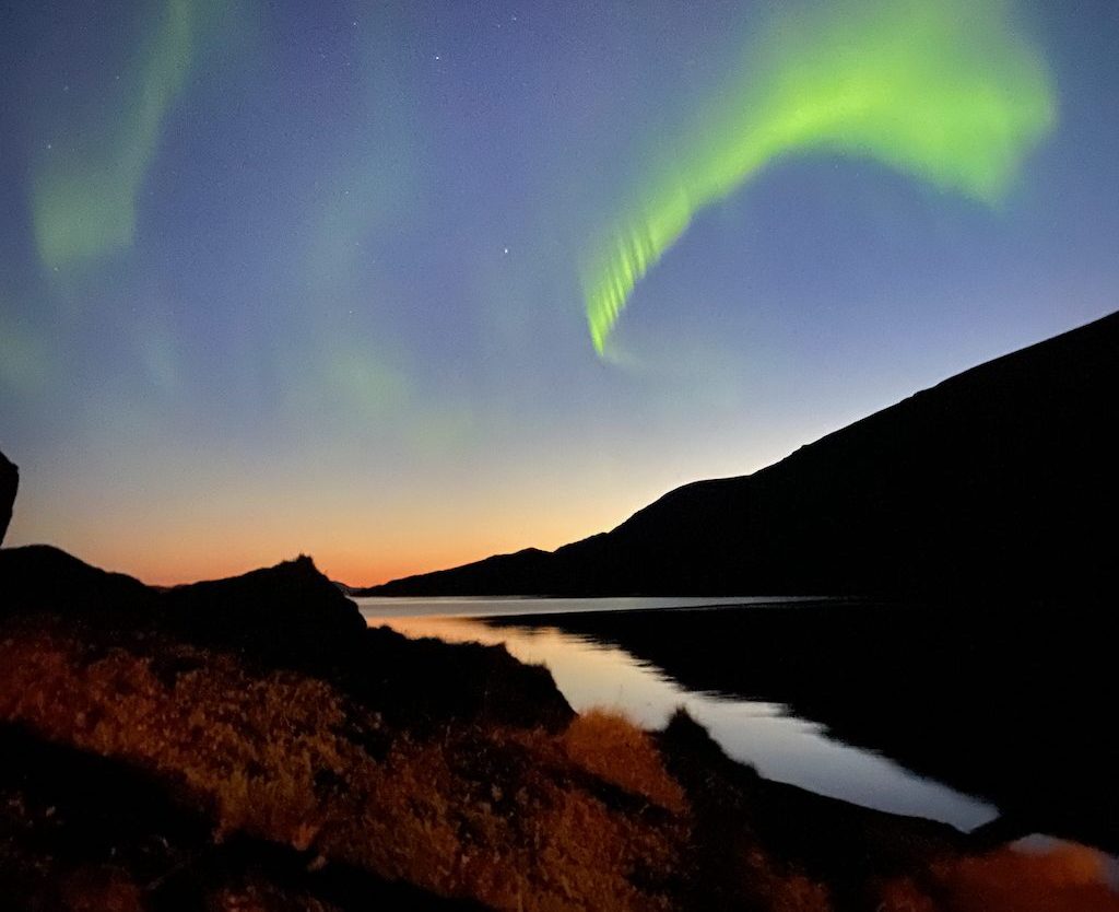 A breathtaking view of the Northern Lights (Aurora Borealis) dancing across the night sky above a calm fjord. The green lights contrast against the deep blue twilight, reflecting beautifully on the still water, with silhouetted mountains framing the scene.
