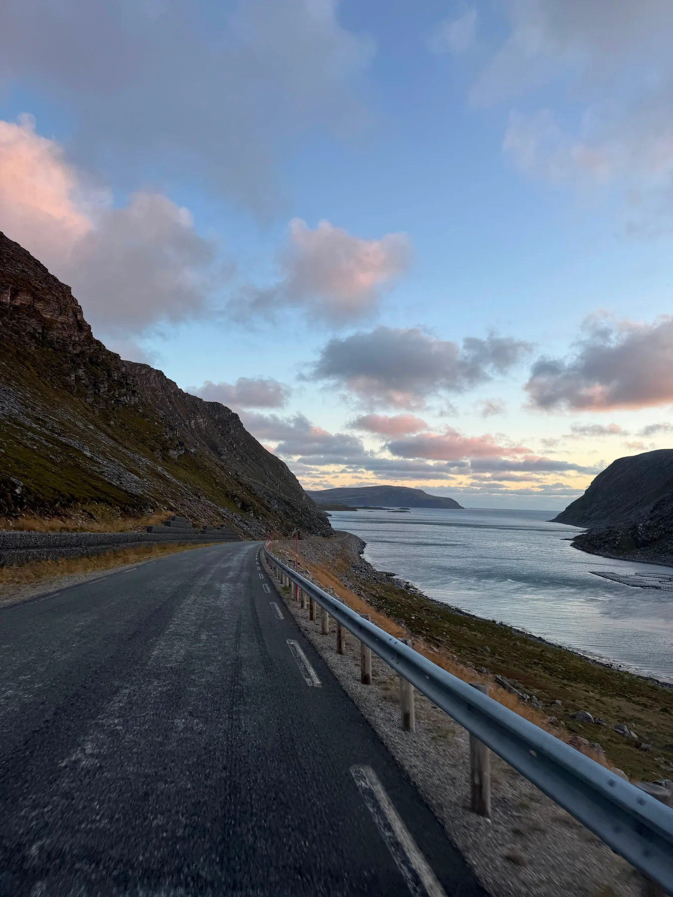 A road follows the rocky fjord shore beneath a softly coloured evening sky.