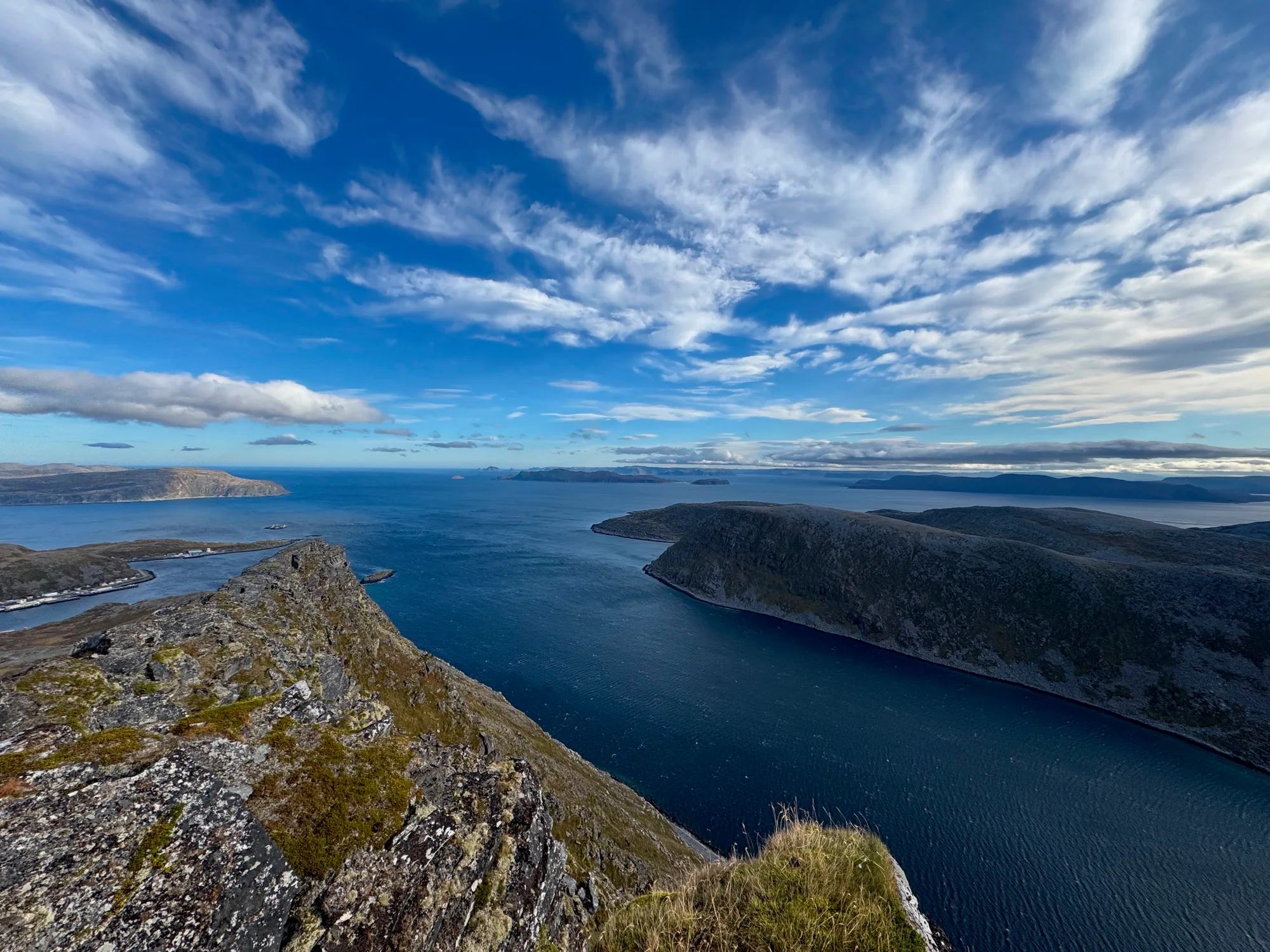 A wide fjord and rocky islands beneath a blue sky, seen from a mountain ridge.