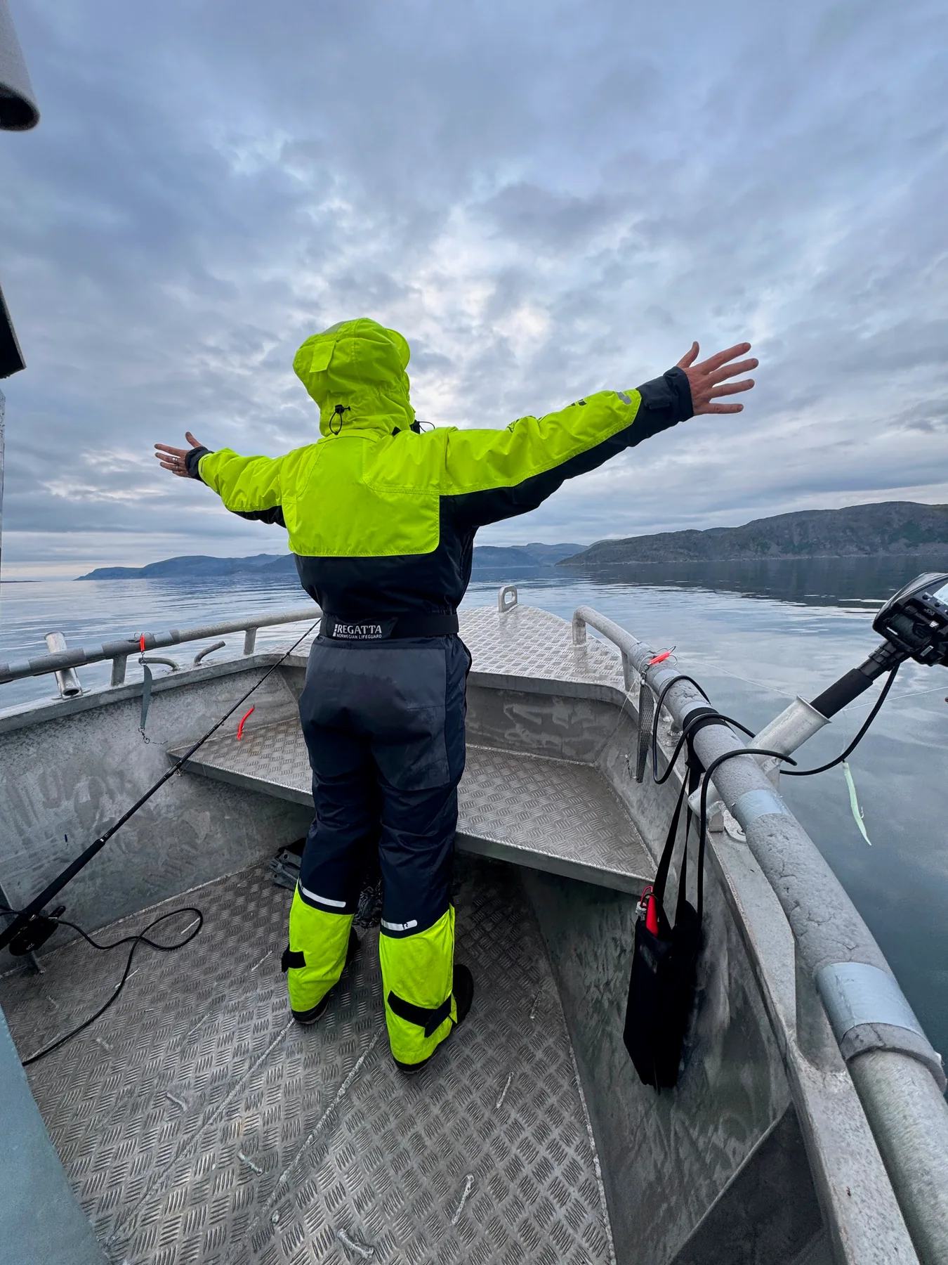 An angler in a yellow flotation suit stands on an open deck, arms outstretched towards the fjord.