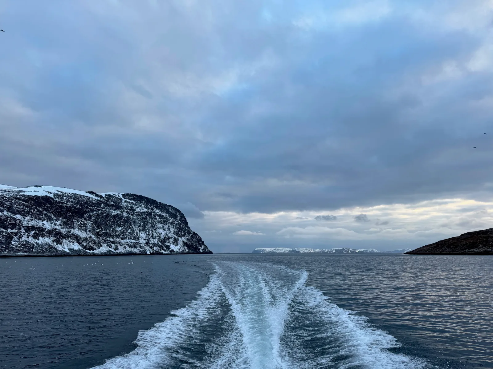 A boat's wake stretches between snow-covered shores under a grey sky.