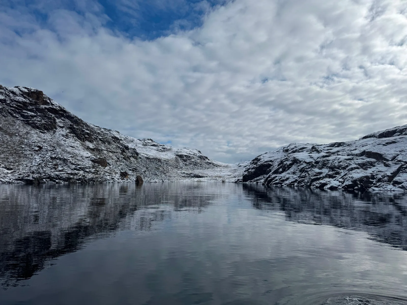 Snow-covered hills reflected in the calm water of a narrow fjord.