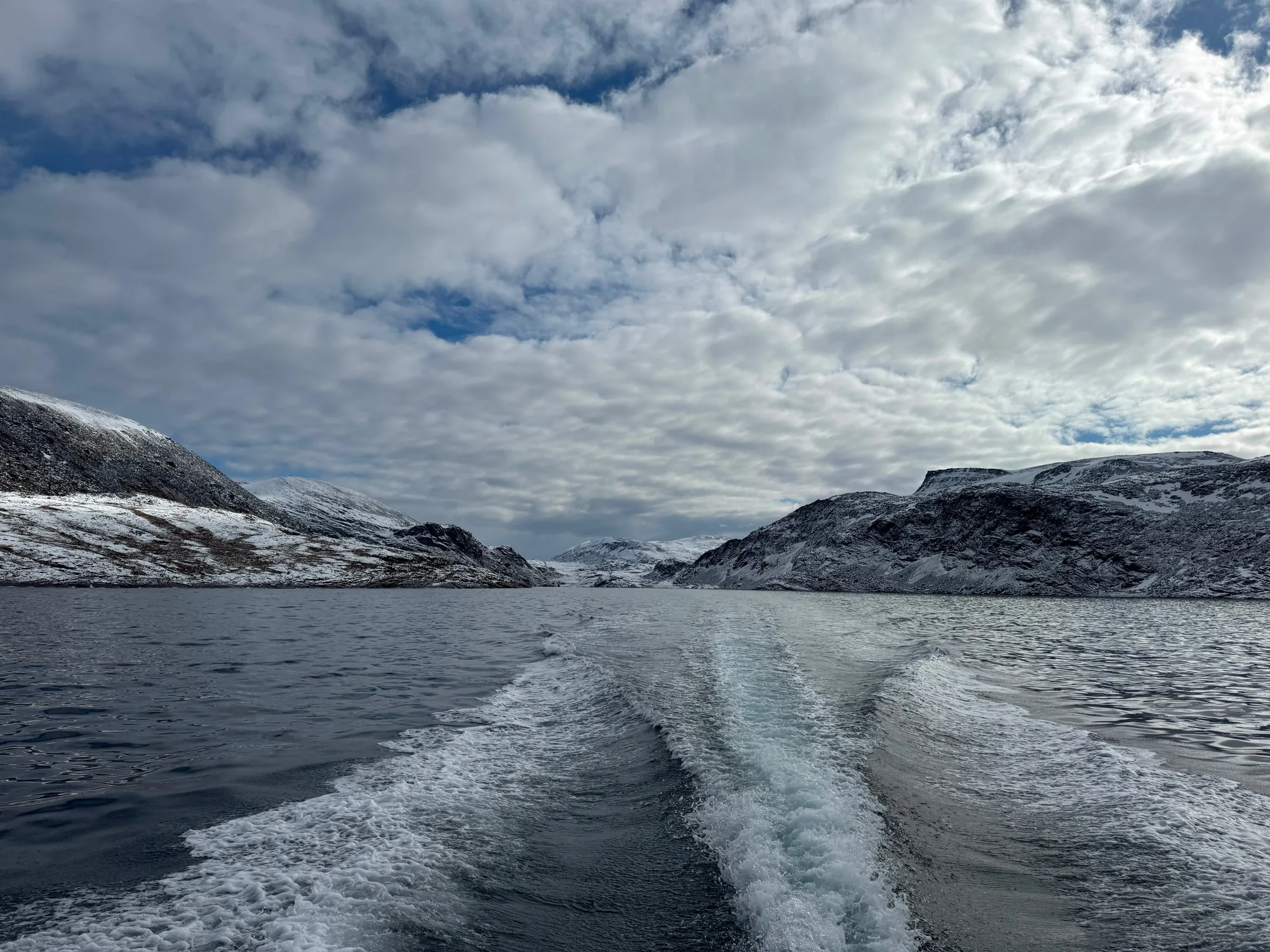 The wake of a boat leads between snow-covered rocky shores beneath a cloudy sky.